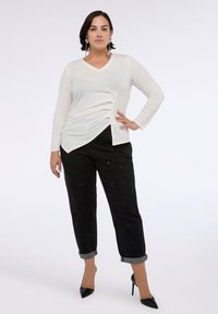 Top bianco a maniche lunghe con scollo a V e un accento drappeggiato, abbinato a jeans neri a tre quarti con dettagli ricamati e risvolti grigi.