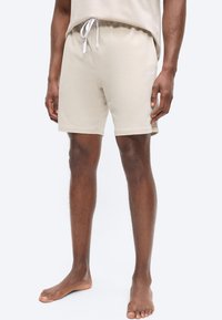 Άνδρας που φοράει μπεζ σορτς με κορδόνι και το αντίστοιχο τοπ, στέκεται ξυπόλητος μπροστά σε απλό ανοιχτό φόντο.