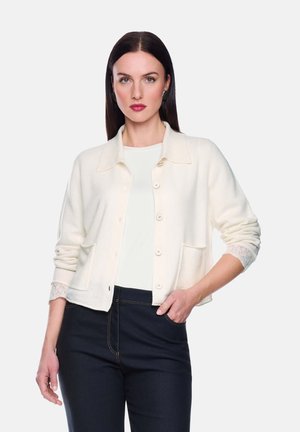 Hellbeige Strickjacke aus weichem Stoff, mit Kragen, durchgehender Knopfleiste, zwei Taschen und Spitzenakzenten an den Ärmeln. Kombiniert mit dunklen Jeans.