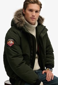 Veste matelassée vert foncé avec une capuche en fausse fourrure, fermeture éclair et logo Superdry Everest sur le bras gauche. Superposée avec un pull en tricot à mailles torsadées crème.