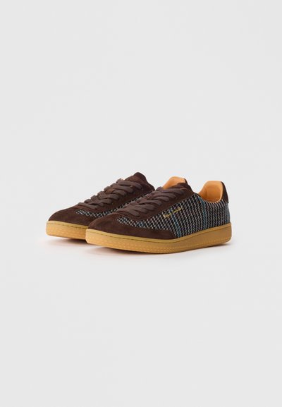 Brūno zamšsneakers ar rakstainu audumu un gumijas zoli, tumšbrūnām šņorēm, novietoti uz vienkārša balta fona.