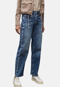 Blauwe denim relaxte-fit jeans met een hoge taille, voorzien van contrasterende stiksels en zijzakken, gecombineerd met witte sneakers en een beige jas.