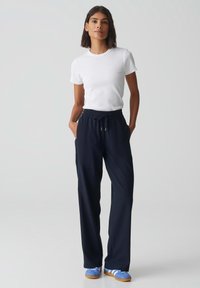T-shirt blanc à manches courtes associé à un pantalon bleu marine à coupe ample avec une taille élastique. Le modèle porte des chaussures bleues avec des rayures blanches.