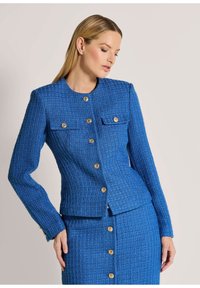 Blauwe textuurtje jas met lange mouwen, nauwsluitend ontwerp en gouden knopen. Heeft twee borstzakken en een ronde halslijn.