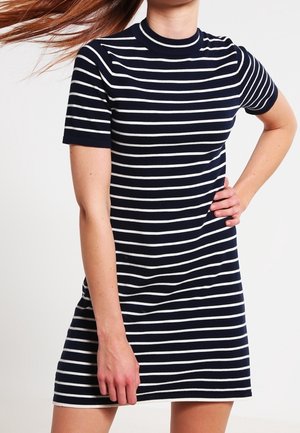 Donna che indossa un vestito blu navy a maniche corte con strisce orizzontali bianche, in piedi con una mano sui fianchi.
