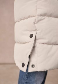 Helle beige Puffjacke mit gestepptem Design, ausgestattet mit dunklen Knöpfen und einem Druckknopfverschluss am Saum. Der Stoff ist glatt und leicht glänzend.
