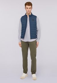 Námořnická puffer vesta s vertikálním prošíváním, nošena přes světle šedou košili. Olivově zelené kalhoty a bílé tenisky dotvářejí outfit.