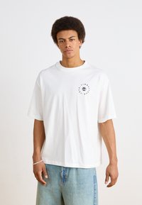Camiseta de algodón blanca con cuello redondo y mangas cortas. Presenta un estampado gráfico negro en el lado izquierdo del pecho. Diseño de corte suelto.
