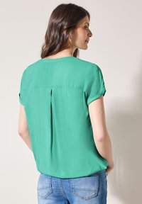 Kortærmet grøn bluse lavet af letvægtsstof, med en rygpilesøm og opsmøgte ærmer, parret med denimjeans.