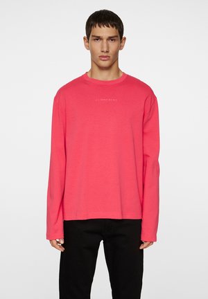 Langærmet T-shirt i lys pink bomuld, med rund hals og subtilt hvidt logo centreret på brystet. Glat tekstur, afslappet pasform.