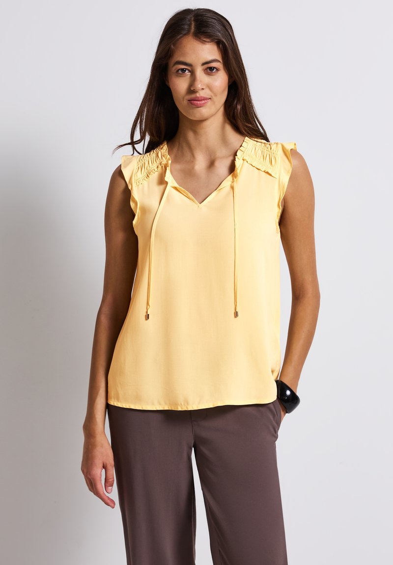 Blouse sans manches jaune avec un décolleté en V, accents froncés aux épaules et cordons de serrage réglables au niveau du col. Tissu lisse, coupe décontractée.