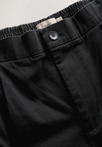 Black cotton pants with an elastic waistband, a single black button fastener, and white label tag visible at the top. Smooth texture.