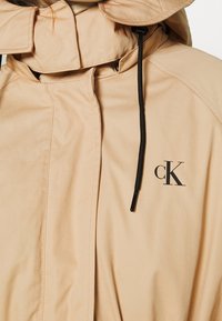 Light beige hooded jacket with a zip closure, featuring a drawstring and a small black 'ck' logo on the front. Smooth fabric texture.
