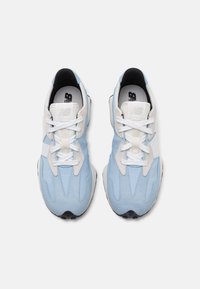 Ett par ljusblå och vita New Balance-sneakers med vita snören, sett ovanifrån på en enfärgad vit bakgrund.