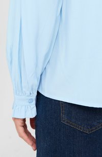 Hellblaue Bluse mit ballonartigen Ärmeln, gefalteten Manschetten und Knopfdetails. Kombiniert mit dunkelblauen Jeans aus Denim mit gestepptem Gesäßtaschen.