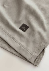 Tissu gris clair perforé avec un patch carré noir comportant un logo triangulaire géométrique blanc près de la couture et de l'ourlet.