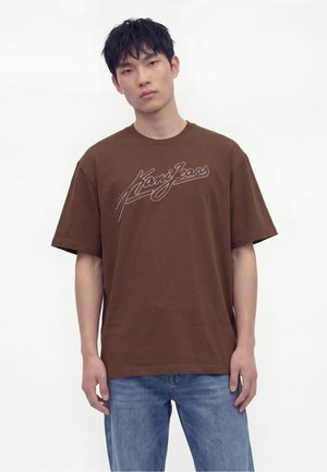 Junger Mann mit kurzem dunklem Haar steht nach vorne gerichtet, trägt ein braunes "Kani Jeans" T-Shirt und hellblaue Jeans vor einfachem Hintergrund.