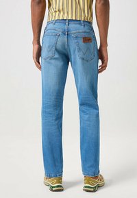 Persona che indossa jeans a gamba dritta blu chiaro con tasche posteriori e scarpe da trekking marroni, in piedi contro uno sfondo chiaro e semplice.