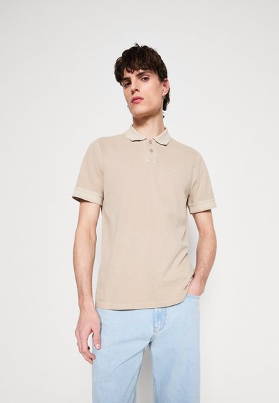 Polo beige en coton avec un col, une patte de boutonnage à deux boutons, et des manches courtes. Détail du logo sur la poitrine. Associé avec un jean bleu clair.