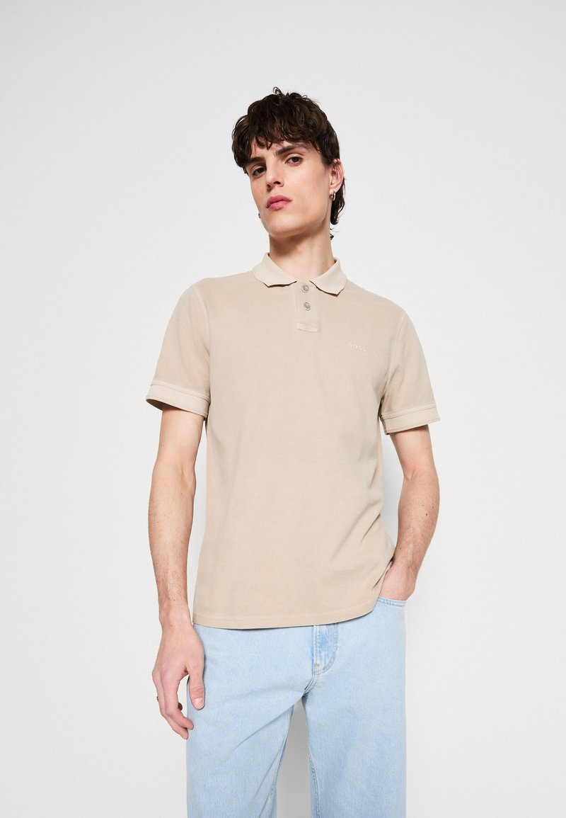 Polo beige de algodón con collar, tapeta de dos botones y mangas cortas. Detalle del logo en el pecho. Combinado con unos vaqueros azul claro.