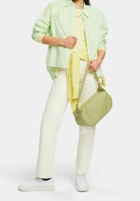 Femme portant une chemise rayée vert clair, un pull jaune noué à la taille, un pantalon blanc, des baskets blanches, tenant un sac à main vert clair.