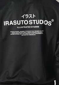 Garbido in tessuto nero con testo bianco: "IRASUTO STUDIOS" e "ILLUSTRATED STUDIOS," con caratteri giapponesi sopra. Il testo è in grassetto e chiaro.