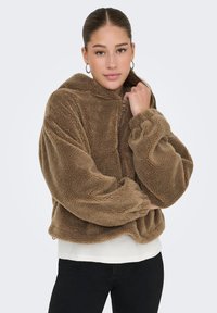 Bruine fleecejas met een hoge kraag, ritsdetail en oversized mouwen. Zachte textuur met een gezellige, casual uitstraling. Draag je over een witte top.