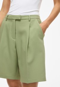 Pantalones cortos entallados de color verde claro con pliegues, bolsillos laterales y una cinturilla con pasadores. Fabricados con una tela suave y de corte estructurado.