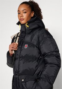 Black padded jacket with a high collar, featuring yellow drawstrings, snap buttons, and an emblem on the chest. Textured finish, casual design.