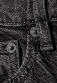 Schwarzer Denim-Stoff mit sichtbaren Nähten, zwei schwarzen Metallknöpfen mit der Gravur "WEEKDAY" und einer Gürtelschlaufe oben.