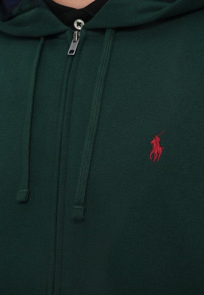 Tmavozelená mikina na zips vyrobená z príjemného materiálu, s malým červeným logom polo na ľavej hrudi a štruktúrovanými šnúrkami.