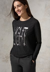 Schwarzes Langarm-Top mit silbernem Grafiktext "LIGHT", aus strukturiertem Stoff, kombiniert mit dunklen Hosen mit gebundenem Bund.