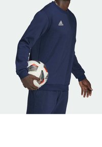 Felpa atletica a maniche lunghe di colore navy con scollatura rotonda, pantaloni abbinati e logo Adidas. La persona tiene un pallone da calcio a strisce bianche e arancioni.