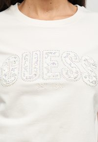 Λευκό μπλουζάκι με τη λέξη "GUESS" να σχηματίζεται από στρας και το "EST. 1981" κεντημένο κάτω από την περιοχή του στήθους.