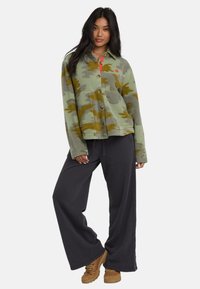 Donna che indossa una giacca in pile verde e marrone camouflage, pantaloni neri larghi e stivali da trekking beige, in piedi con una postura rilassata su sfondo bianco.