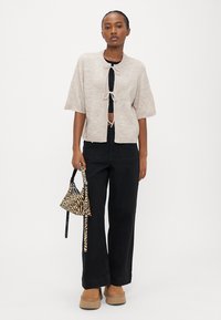 Woman wearing beige short-sleeve cardigan tied in front, black wide-leg pants, tan platform shoes, holding a small zebra print handbag.