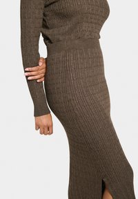 Robe tricotée en marron foncé avec une texture côtelée, des manches longues et une fente sur le côté. Silhouette ajustée qui met en valeur la forme du corps.