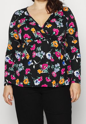 Schwarzes Wickeloberteil mit langen Ärmeln und rosa, orangefarbenem, weißem und blauem Blumenmuster, getragen zu schwarzen Hosen an einer Frau.