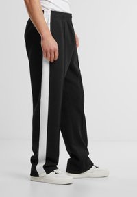 Sorte sweatpants med en hvid sidelæbe, der har en elastisk talje og en lige benlængde, parret med hvide sneakers.