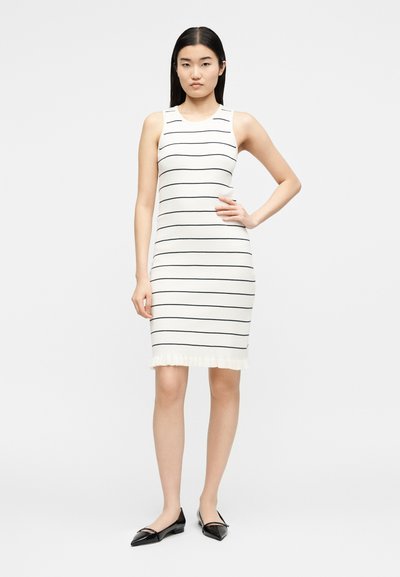 Γυναίκα στέκεται με το αριστερό χέρι στη μέση φορώντας λευκό αμάνικο φόρεμα με λεπτές μαύρες οριζόντιες ρίγες και μαύρες μυτερές φλατ γόβες.