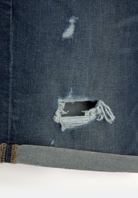 Tissu en denim de couleur bleu foncé, présentant des zones usées et une grande déchirure. L'ourlet est fini dans une nuance de bleu plus clair.