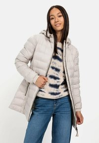 Donzen jas in lichtbeige met een capuchon, met een doorgestikt ontwerp en een rits aan de voorkant. Draag over een crèmekleurige sweater met blauwe patronen en denim jeans.