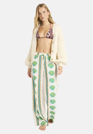 Gebreid crèmekleurig vest met oversized mouwen, gedragen over een bordeauxrode bikini met patroon en crème broek met groene schelpmotieven en verticale strepen.