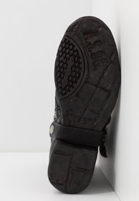 Svart gummisula med texturerat greppmönster, böjd form, och ett band med en dekorativ metallknapp. Storleksmärkning synlig.