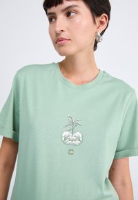 Mintgrön kortärmad t-shirt i bomull med en grafisk tryck av händer som håller en liten planta. Enkel rund hals och mjuk textur.
