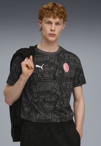 Sort T-shirt med korte ærmer med gentagne tekstdesigns, en hvid Puma-logo og et rødt AC Milan-våbenskjold, båret med en sort jakke over skulderen.