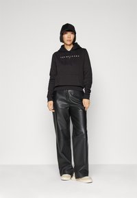Sudadera negra con capucha y logo, combinada con pantalones de cuero negros de cintura alta y zapatillas beige; presenta un corte relajado y un estilo casual.