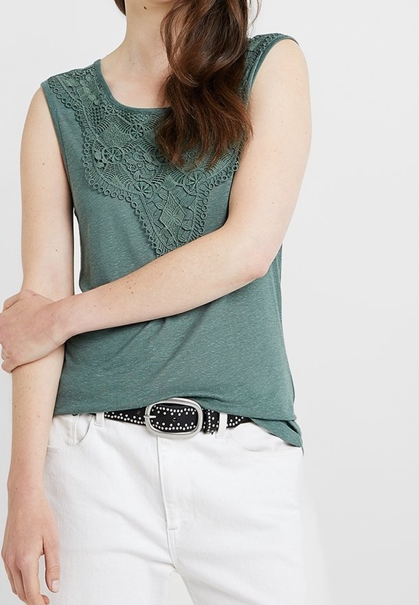 Zielona bezrękawnik z misternym koronkowym wykończeniem z przodu, połączona z białymi spodniami i czarnym paskiem z srebrnymi akcentami.