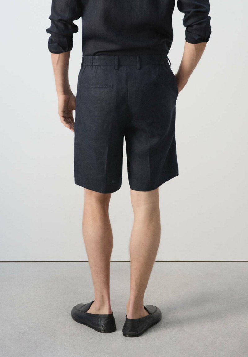 Homme portant une chemise noire à manches longues, un short noir avec passants pour ceinture et des chaussures noires à enfiler, debout sur un sol gris contre un mur blanc.