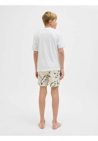 T-shirt en coton blanc associé à un short beige avec un motif abstrait noir et vert, vu de dos. Pieds nus.